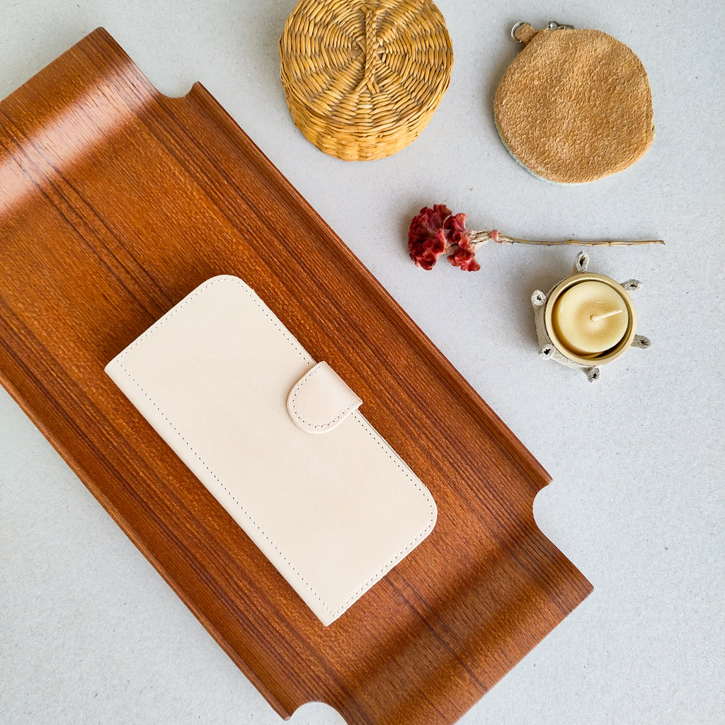 A flat lay of an ivory eco-leather Fairphone case lying on a mid-century vintage brown wooden tray, a white leather star-shaped candle holder, a red dried flower, a small basket with a lid, and a leather circle shaped pouch, on a grey background