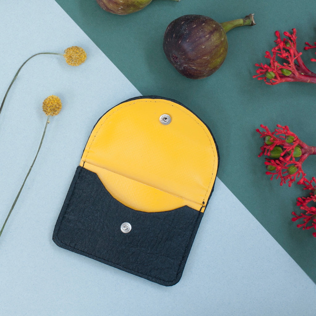 A black vegan wallet made of pineapple leave fibre and yellow waste material lining. In a flat lay composition with figs and flowers